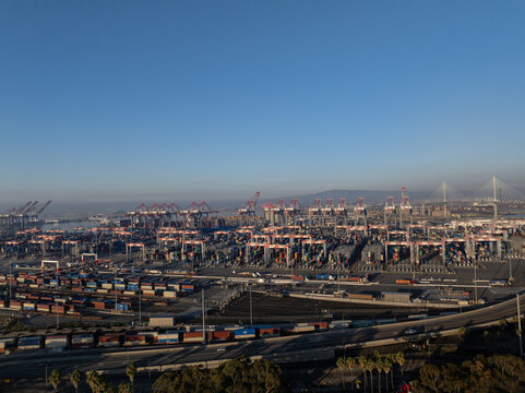 Aerial view of a sprawling port with towering cranes and stacks of colorful containers under a clear sky, Vincent Thomas Bridge in the distance, Los Angeles, California, United States.