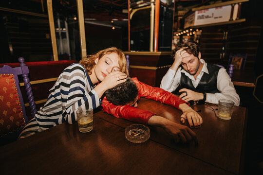 Three friends sit around a wooden table in a dim bar. One woman rests her head on her hand, a man in red leans forward, and another looks stressed, with drinks and an ashtray on the table.