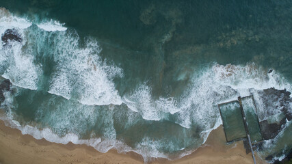 Aerial view of foamy waves crashing onto the sandy shore near the tranquil rock pool, a contrast of wild ocean and serene enclosure, Wollongong, New South Wales, Australia.