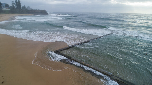 Aerial view of the ocean's restless waves crashing against the sandy shore and the tranquil rock pool, Wollongong, New South Wales, Australia.