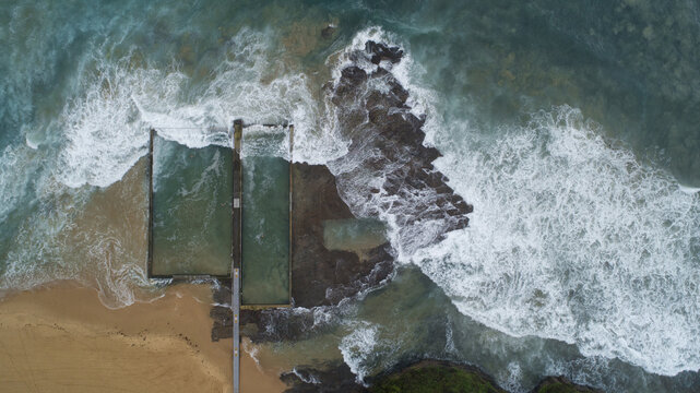 Aerial view of ocean pool embraced by foamy waves against the rocky coast and sandy beach, Wollongong, New South Wales, Australia.