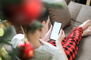 Over-the-shoulder shot of boy in red pajamas lying on sofa using smartphone with blank white screen...