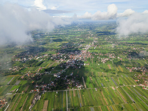 Aerial view of a vibrant tapestry of green rice fields and terracotta rooftops nestled under a blanket of clouds, Klaten Regency, Central Java, Indonesia.