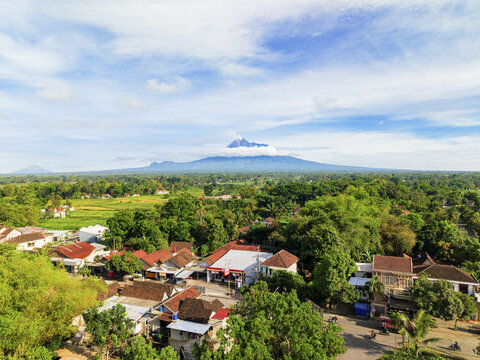 Aerial view of the lush green landscape dotted with buildings, stretching towards a majestic mountain under a sky, Klaten Regency, Central Java, Indonesia.
