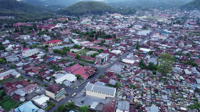 A high-angle aerial drone shot over Bajawa, Flores, Indonesia. The view looks down a long, straight main street, revealing the dense town's red and tin roofs.
