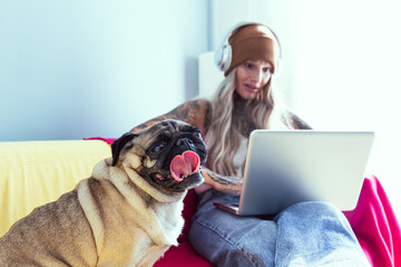 A tattooed woman sits comfortably on a couch, focused on her laptop. A playful pug dog sits next to her, creating a cozy and relaxed atmosphere in a bright room.