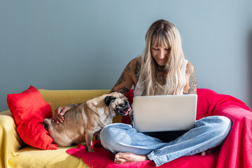 A tattooed woman sits on a colorful couch, focused on her laptop. Her pug dog patiently sits nearby, creating a cozy atmosphere in their home. Natural light brightens the scene.