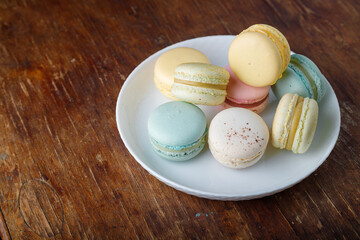 Tender multicolored macaroons with different tastes on white plate, wooden old table background, closeup shot, copy space