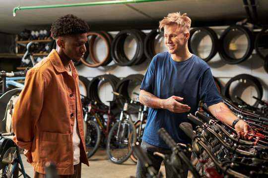 Young man explaining bicycle features to Black young man in bike shop, both standing among rows of bicycles with wheels displayed on wall in background