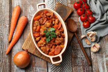 Delicious cooked stew in baking dish, fresh ingredients and spoon on wooden table, flat lay