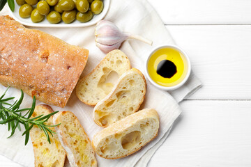 Fresh cut ciabatta, oil, olives, garlic and rosemary on white wooden table, top view. Space for text
