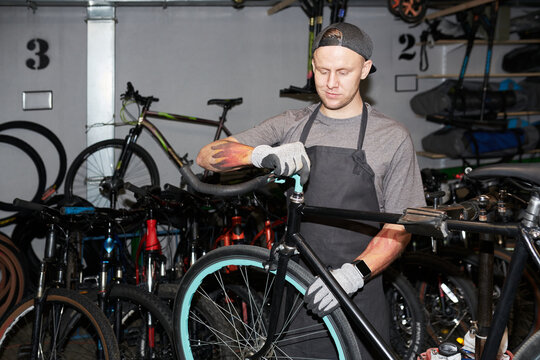 Young man adjusting handlebars on bicycle in workshop, wearing gloves and apron, surrounded by multiple bikes and spare wheels, focused on repair task