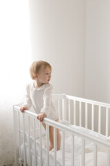 cute smiling healthy baby girl in beige bodysuit is standing near the crib at home in the nursery with teddy bears on white bedding, the child is learning to walk