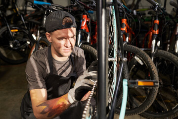Young man wearing cap and gloves repairing bicycle wheel in workshop, focusing on adjusting chain mechanism while surrounded by multiple bikes in background