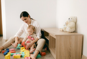 Young happy mother playing game with educational pyramid with cute little child girl in the living room at home. Woman parent spending time with daughter. Family leisure and people concept.