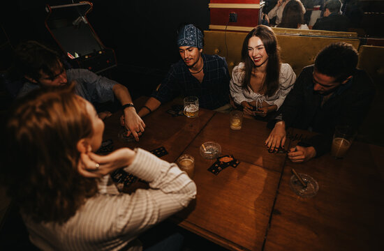 A lively group of friends shares a card game at a dimly lit bar, smiling and chatting as drinks sit on the wooden table.