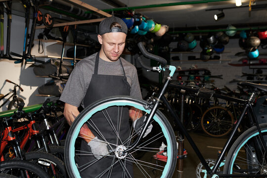 Young man wearing backwards cap repairing bicycle wheel in workshop, standing among various bikes and tools, focusing on maintenance task with hands on tire