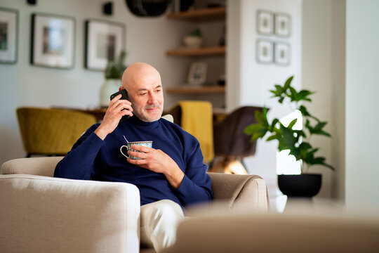 A man relaxing in an armchair in his modern home and using phone and drinking tea