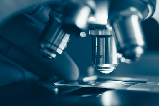 Macro photo of optical microscope lens focusing on sample slide under sterile laboratory lighting. Symbolizes technology, research, and medical discovery.