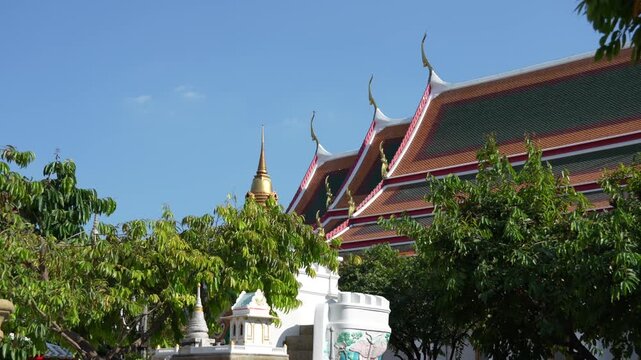 Exterior view of Wat Pho, Bangkok, featuring the gilded Chofa finials and tiered, tiled temple roof, with a smaller golden chedi (stupa) and lush trees beneath a clear blue sky.