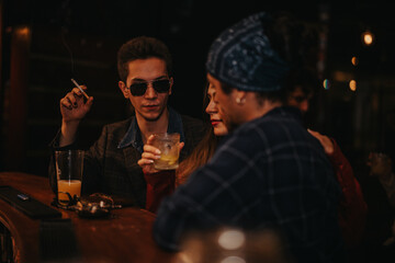 A group of friends relax at a bar as one man smokes and others sip drinks. Moody, intimate lighting and casual urban style create a nightlife vibe.