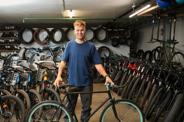Portrait of young man standing in bicycle workshop holding bicycle, surrounded by rows of bikes and spare tires, smiling at camera, short blond hair visible