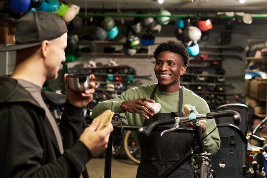 Young Black man wearing apron standing in bicycle workshop smiling and holding sandwich while talking to young man holding mug and sandwich, helmets and bikes visible in background - Powered by Adobe