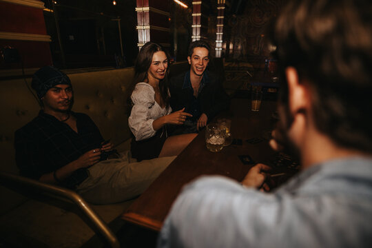 A small group of friends sit around a wooden table in a moody venue, drinking and chatting while playing cards. Warm lighting and casual conversation convey relaxed nightlife and companionship.