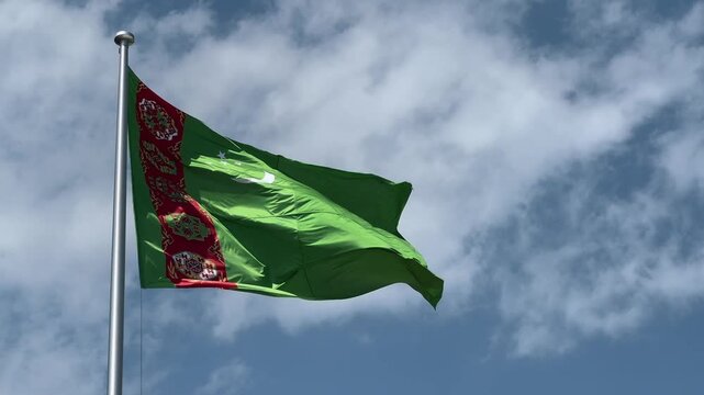 A close-up shot of the flag of Turkmenistan fluttering vibrantly in the wind under a bright sky, showcasing its rich green color and intricate carpet pattern in stunning detail.