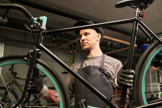 Young man wearing cap and gloves repairing bicycle in workshop, holding bike frame while inspecting components, standing in industrial setting with focused expression