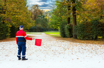 Event supervisor wearing uniform manages outdoor marathon signaling with bright red flag across peaceful autumn park landscape.