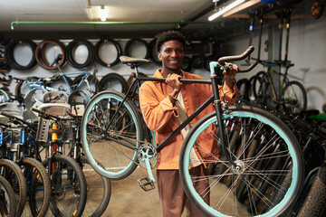 Young Black man smiling while holding bicycle on shoulder in workshop surrounded by various bikes and spare tires, standing confidently and looking at camera