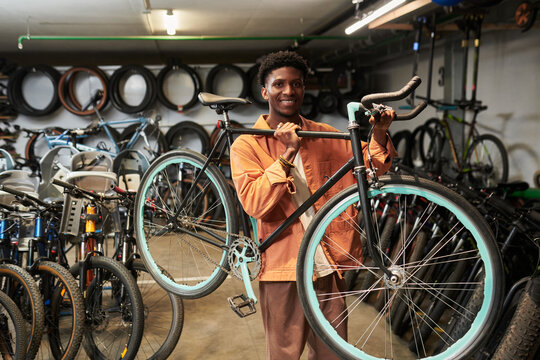 Young Black man smiling while holding bicycle on shoulder in workshop surrounded by various bikes and spare tires, standing confidently and looking at camera