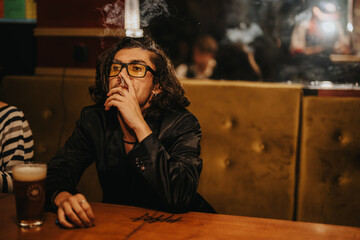 A man with glasses sits in a dim, warm bar booth, smoking and enjoying a beer. Moody, stylish...