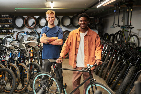 Young man standing with arms crossed next to Black young man holding bicycle, both smiling in bicycle workshop surrounded by various bikes and wheels