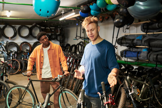 Young man explaining bicycle features to Black young man in bike shop, both standing among multiple bicycles and cycling equipment, engaging in conversation