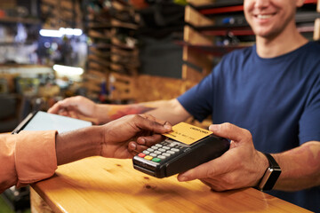 Young man holding payment terminal while Black young man making contactless payment with credit card at counter in retail store, both smiling during transaction