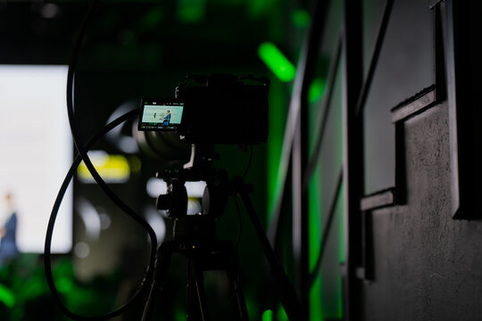 Digital mirrorless camera mounted on tripod filming presentation in conference hall with professional lighting, symbolizing live streaming, event videography and media equipment