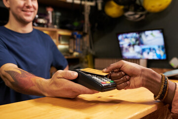 Young man holding payment terminal while Black young woman making contactless payment with credit...