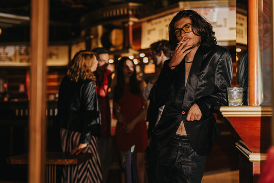 A stylish man in a dark blazer and yellow glasses leans on a bar, smoking, while a blurred group of friends chats in the warm, vintage nightlife setting.