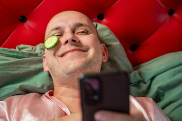 Short-haired man with cucumber slices on face holding smartphone, skincare and relaxation concept