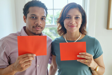 Caucasian woman and Black man standing side by side holding blank red cards in front of chest, both looking at camera with neutral expressions in bright indoor setting