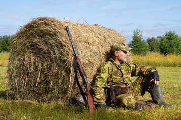 A hunter and his dog relax in the shade of a large round hay bale