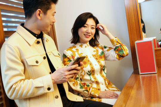 Young adult Asian woman sitting at vanity smiling at camera while young adult Asian man holding smartphone looking at her, both engaging in casual conversation in modern setting