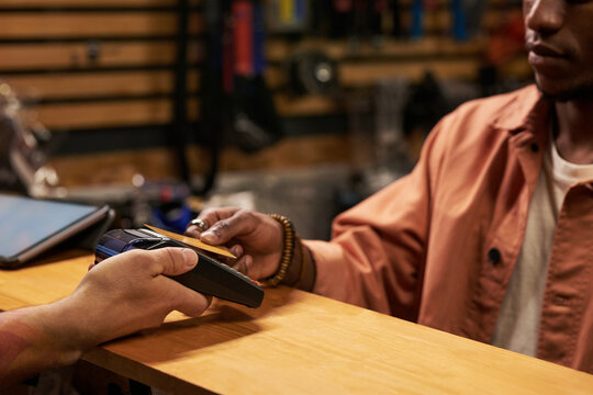 Young Black man making contactless payment with credit card at counter, holding card near payment terminal while another hand presenting device, partial faces visible