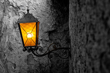 Historic wall lantern inside ancient Predjama castle in Slovenia. The wrought-iron lamp glows with a warm yellow light against dark quarry stone walls (black and white). Atmospheric vintage motif.