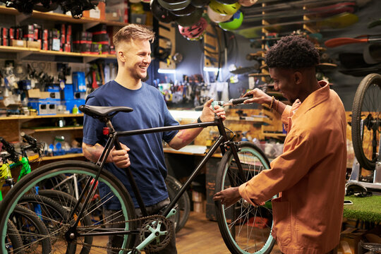 Young man and Black young man working together repairing bicycle in workshop, both standing and smiling while holding bike, tools and helmets visible in background