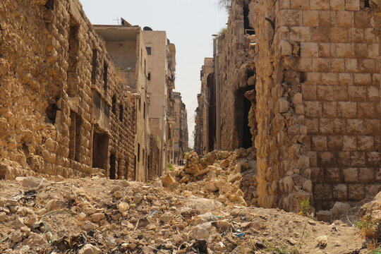 War damaged street in Aleppo, Syria, with destroyed stone buildings and rubble blocking the way. In the distance, people return, symbolizing resilience and the importance of reconstruction.
