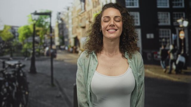 Woman smiling with eyes closed, wearing open shirt and white tank showing bare chest on a street with bicycles and lamp posts; calm contentment.