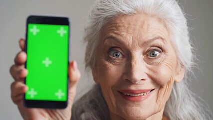 Closeup of a happy senior woman with white hair holding up a smartphone with a green screen and tracking markers looking directly at the camera with a surprised and joyful expression perfect for app .